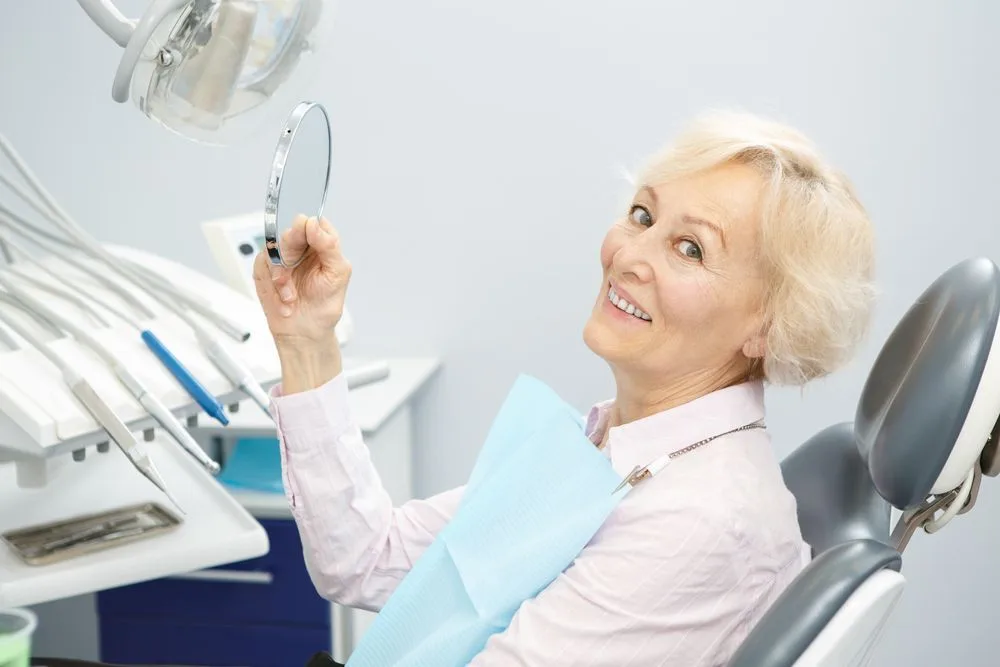 Older patient smiling at her reflection in a hand mirror in the dental chair — dental clinic in Buderim, QLD